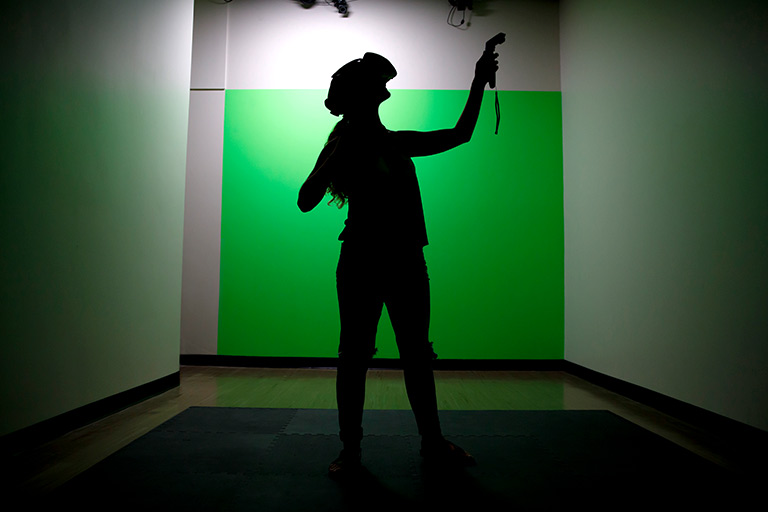 A student stands in front of a green screen with a virtual reality headset and controller.