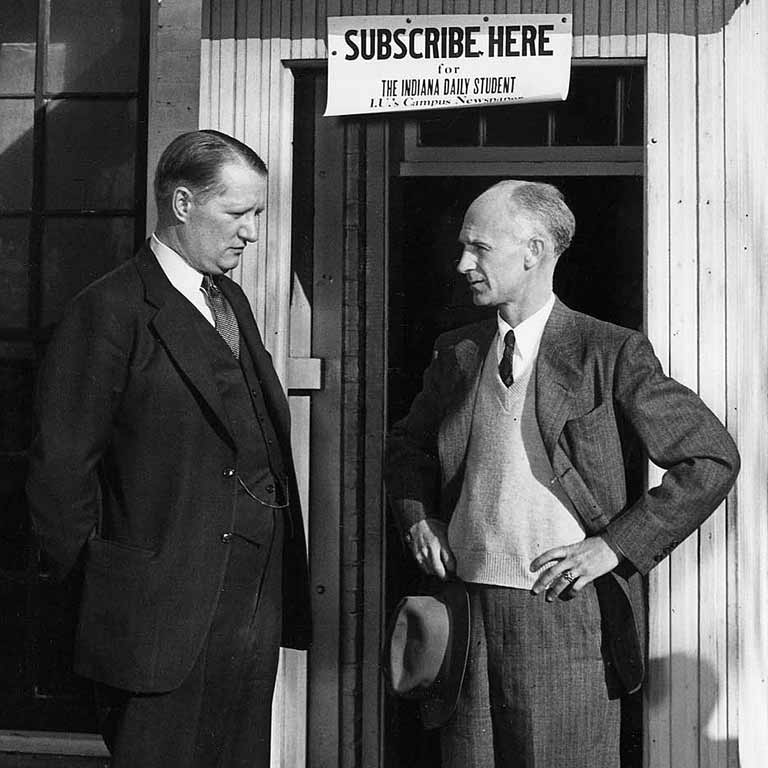 Ernie Pyle talks with a man in a suit in front of a doorway and sign to subscribe to the IDS.