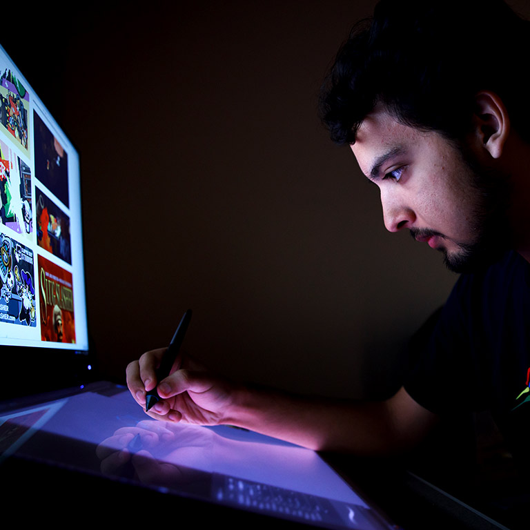 A student draws on a drawing tablet as he looks at a design on a monitor.