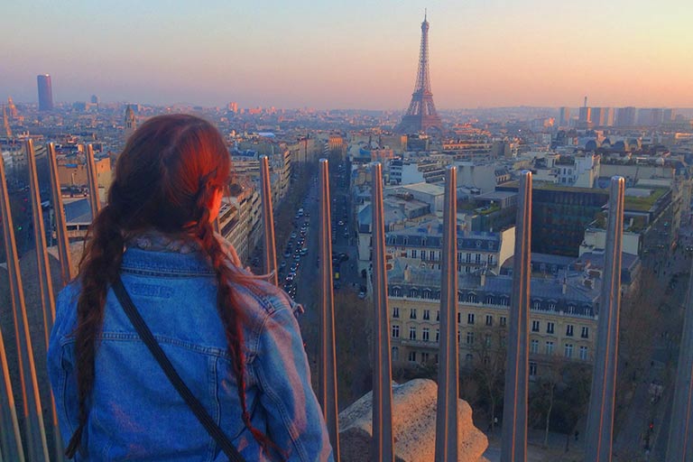 A woman looks out over Paris in the direction of the Eiffel Tower at sunset.