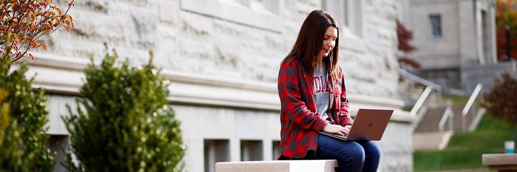 A woman sits on a stone bench outdoors and uses a laptop.