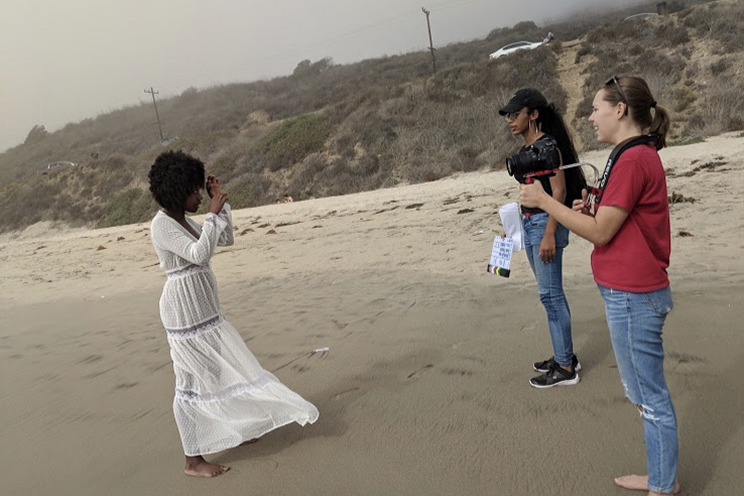 Students stand on a beach with a camera and plan out a shot.