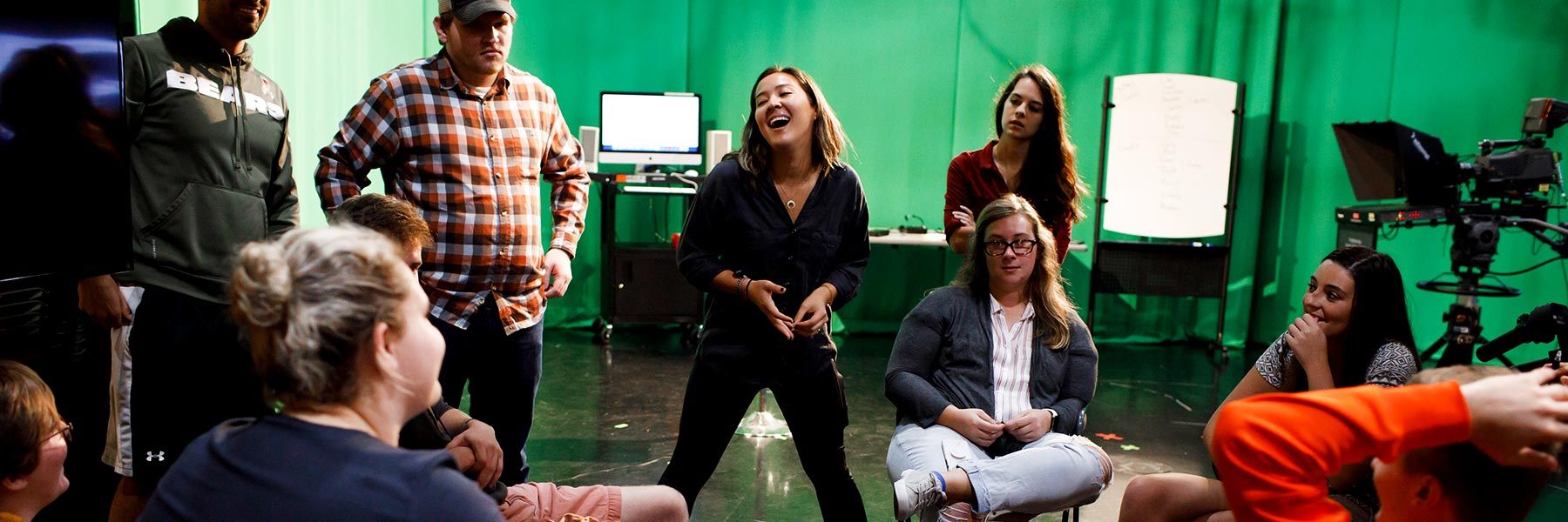 A group of students chat in a circle in a TV studio with a green screen.