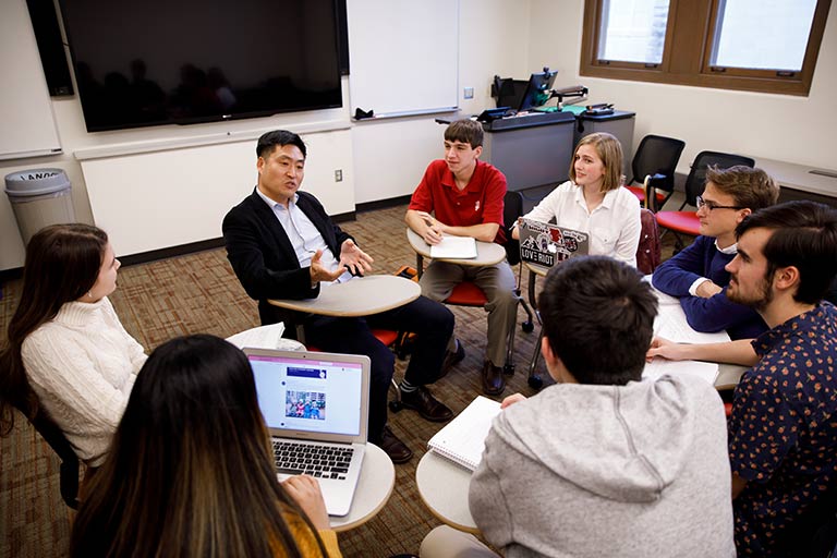 A professor speaks with students as they are seated in a circle in a classroom.