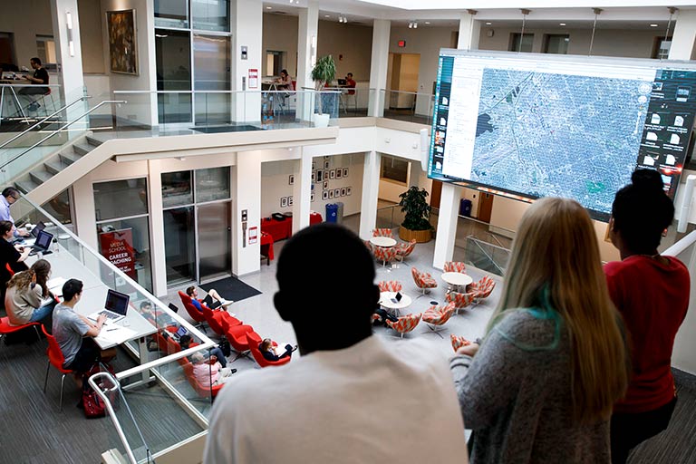 Three students look down at the large screen in the central commons of Franklin Hall from above.