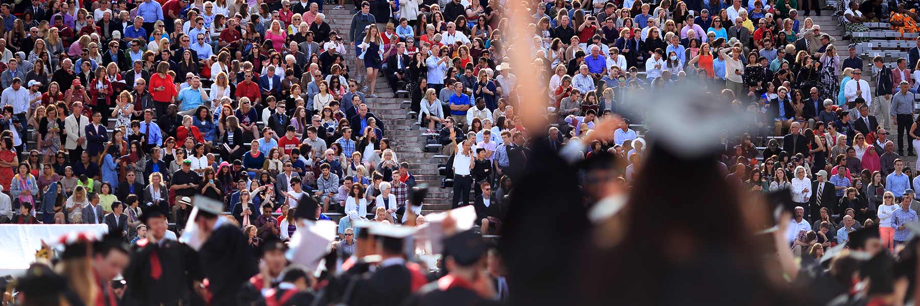 A student reaches toward the sky during graduation as she looks toward the large crowd of attendees.