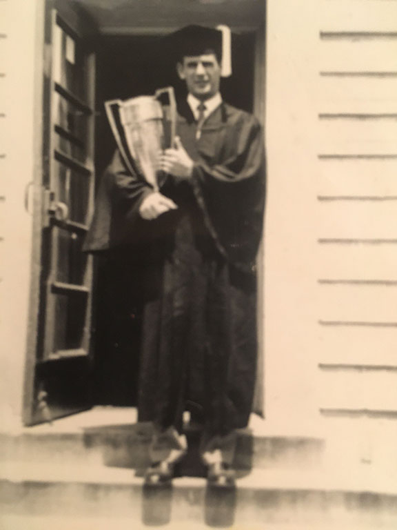 A black and white photo of Bernie Rosenthal standing in a doorway wearing a graduation cap and gown and holding a trophy.