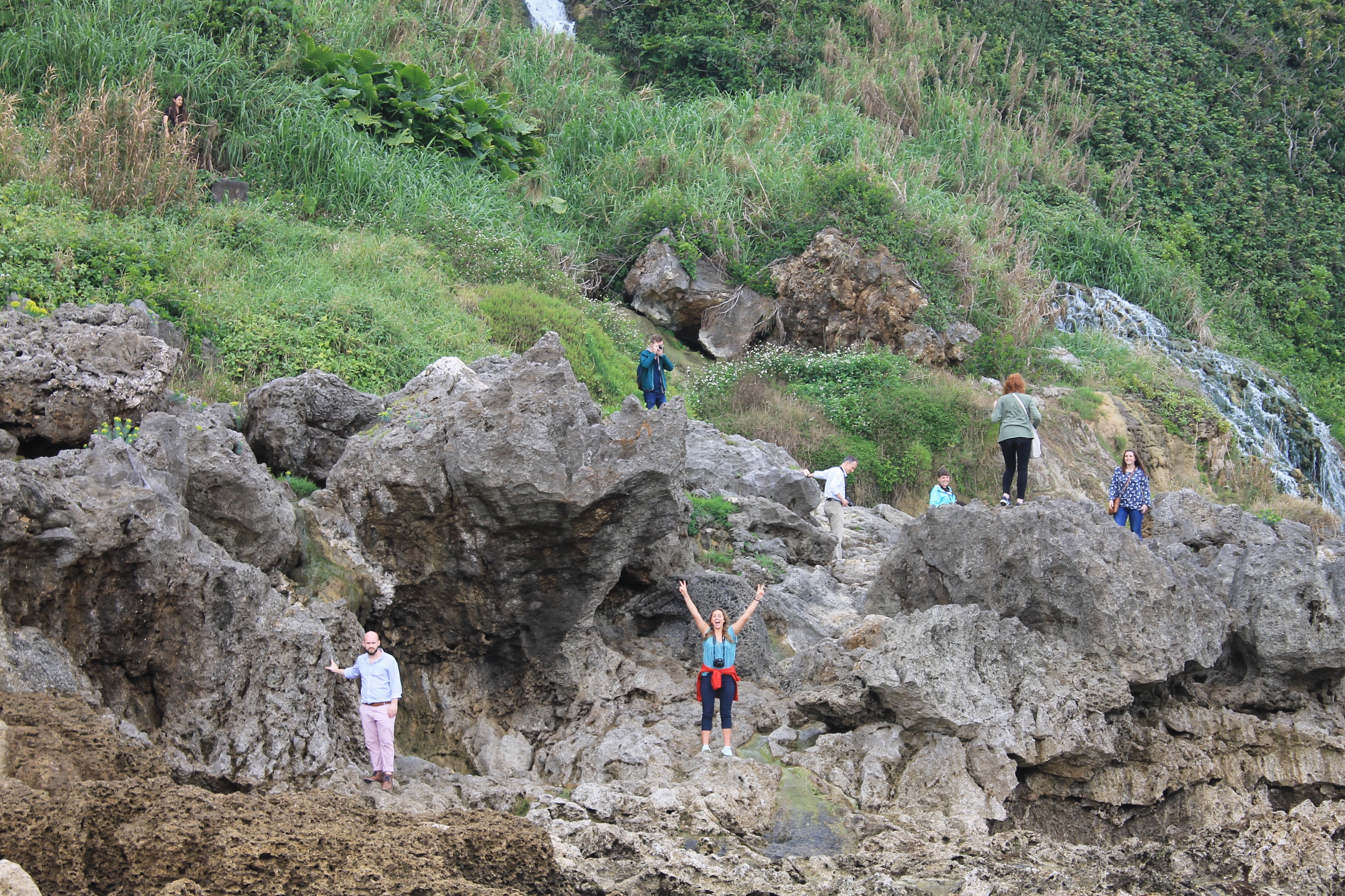 Students climb on a cliff in Okinawa, Japan. One student makes double peace signs with her arms raised.