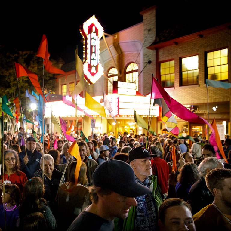 A crowd of people waving colorful flags at night on Kirkwood Avenue outside the Buskirk-Chumley Theater during Lotus fest