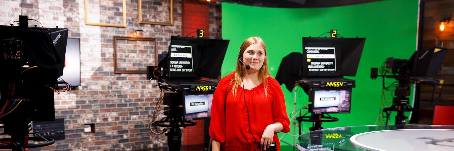 A student with a headset on stands in front of a set of teleprompters in a TV studio.