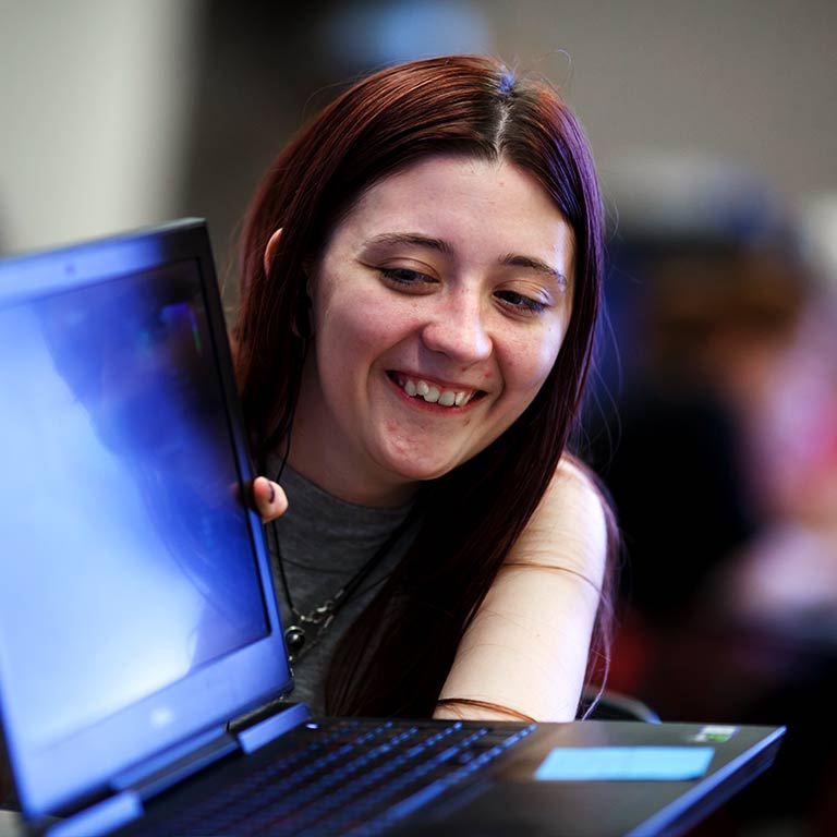 A student smiles and looks around the edge of a laptop screen.