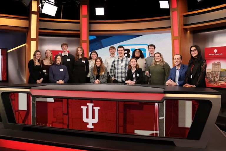 Group of people stand on a TV production set behind a news desk that has the IU logo displayed on front.