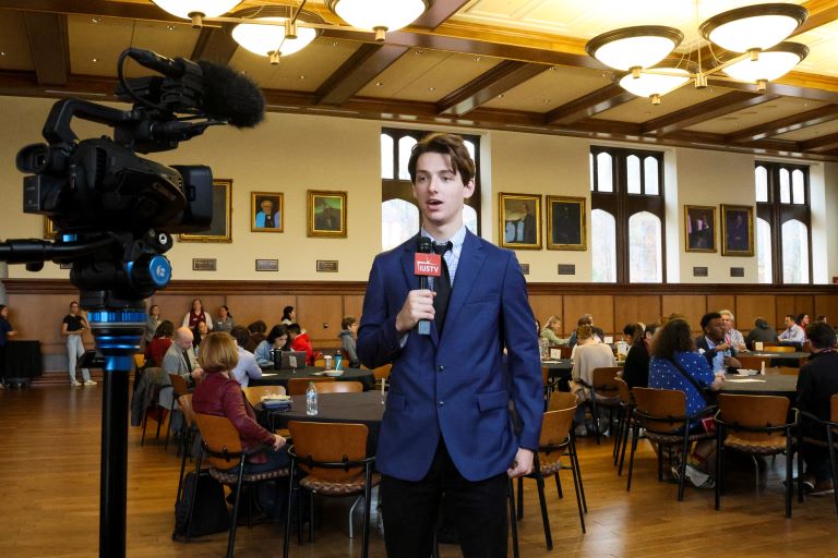 Person holding a microphone speaks toward a camera while a crowd is seated at a variety of tables behind the reporter.