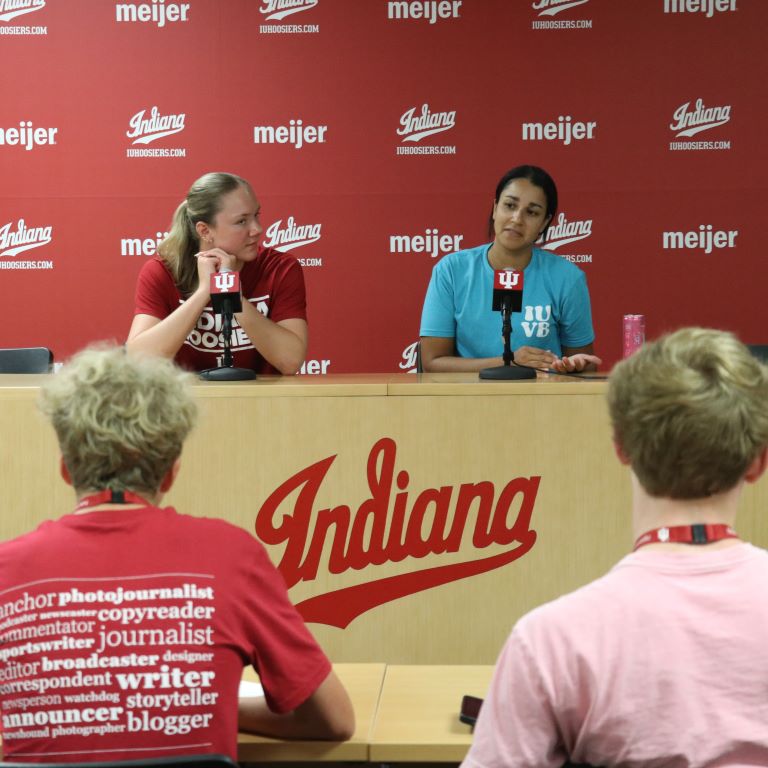 Two people are seated in front of microphones facing two others seated with their backs to the camera.