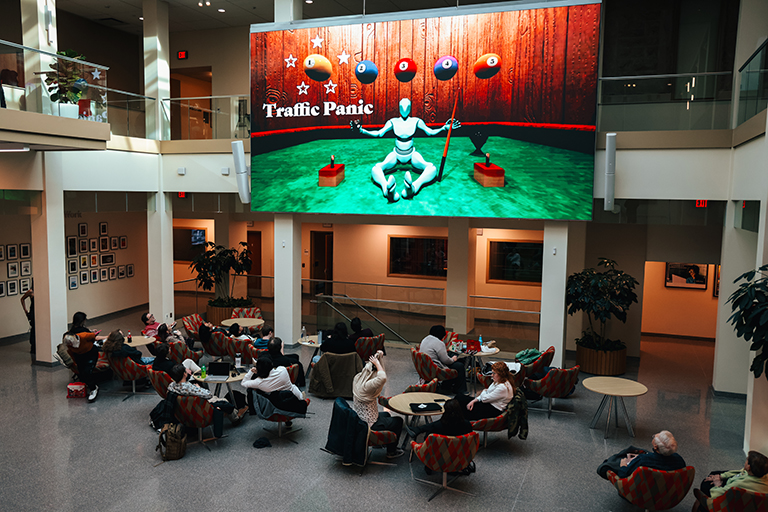 Students sit at tables beneath a large screen showing a mockup of a video game level select menu.