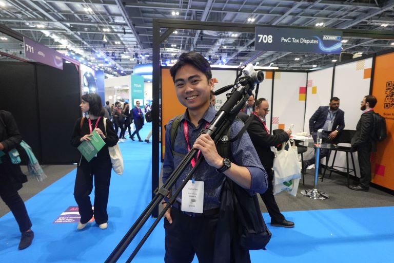 Person holding a camera tripod stands in a large expo room with several company booths filling the area.