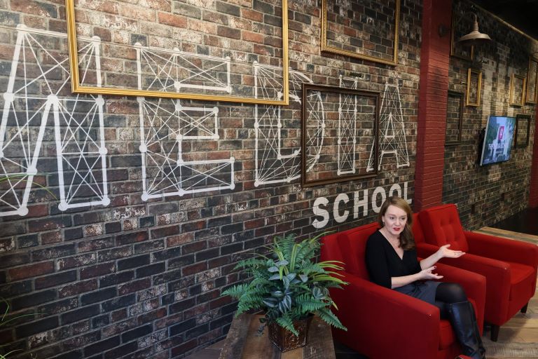 Wide shot of a person sitting in a studio with a brick wall that says "The Media School" displayed behind her.