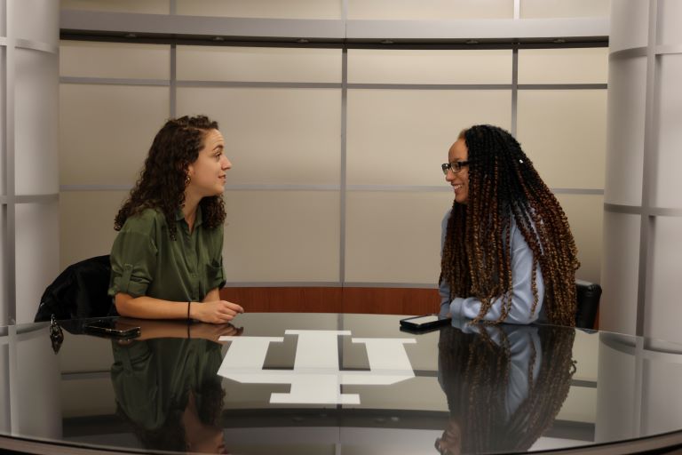 Two people are seated behind a news desk turned towards one another speaking.