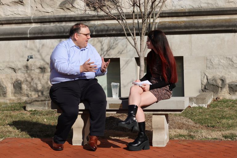 Two people are seated outside on a stone bench talking to one another.