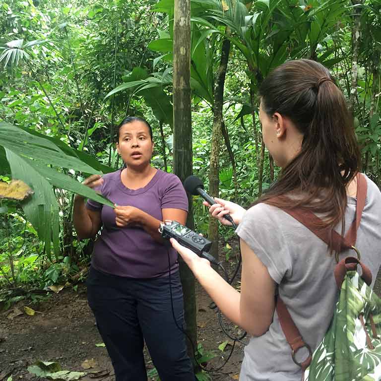 A woman interviews another woman in a rainforest.
