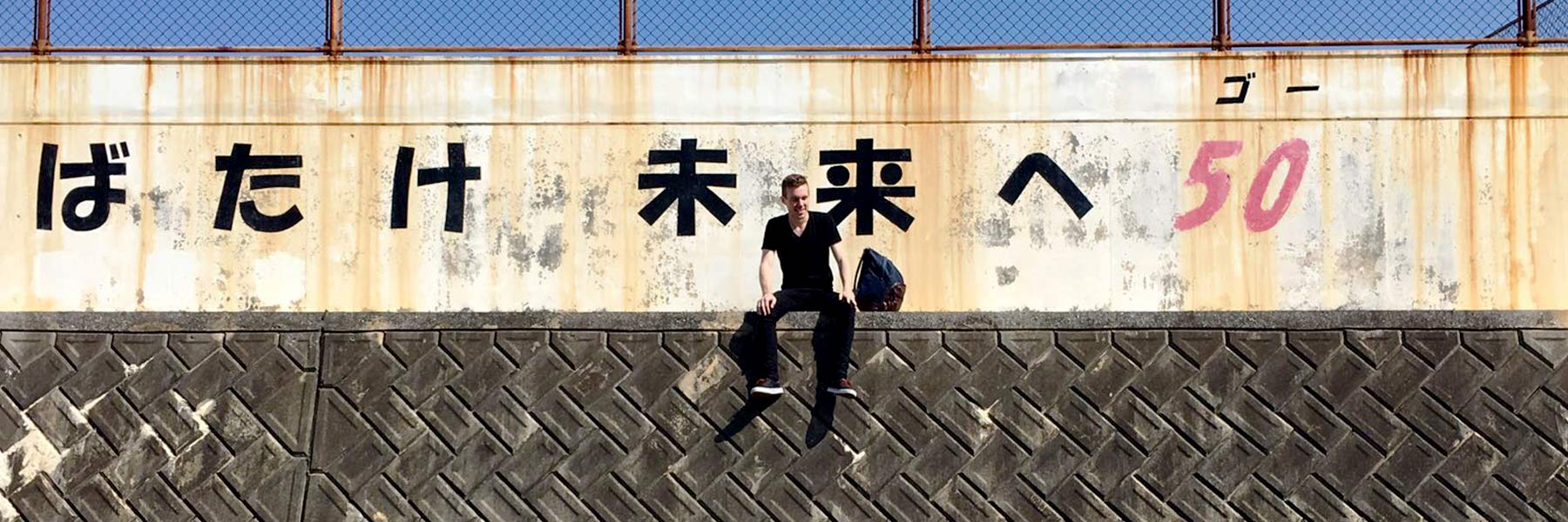 A man dangles his feet over a ledge in front of a wall with Japanese characters on it.