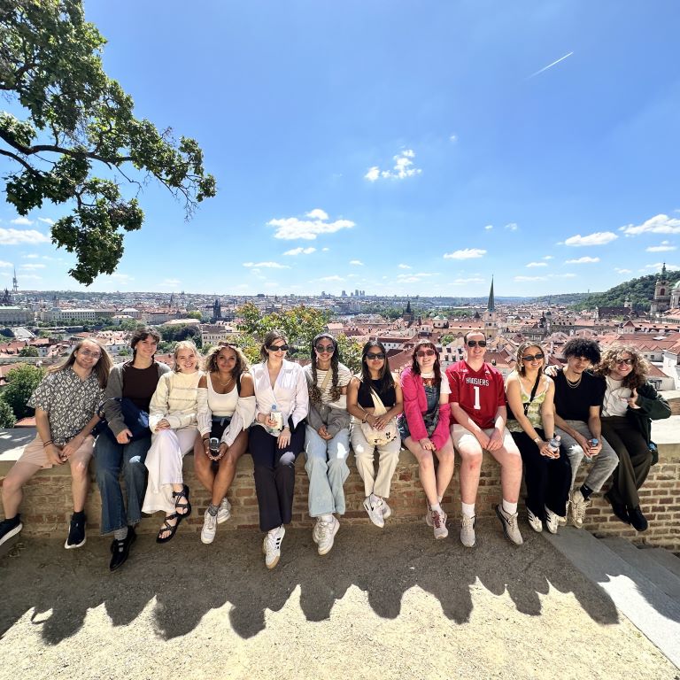 A group of students sit on a stone wall with a cityscape behind them.