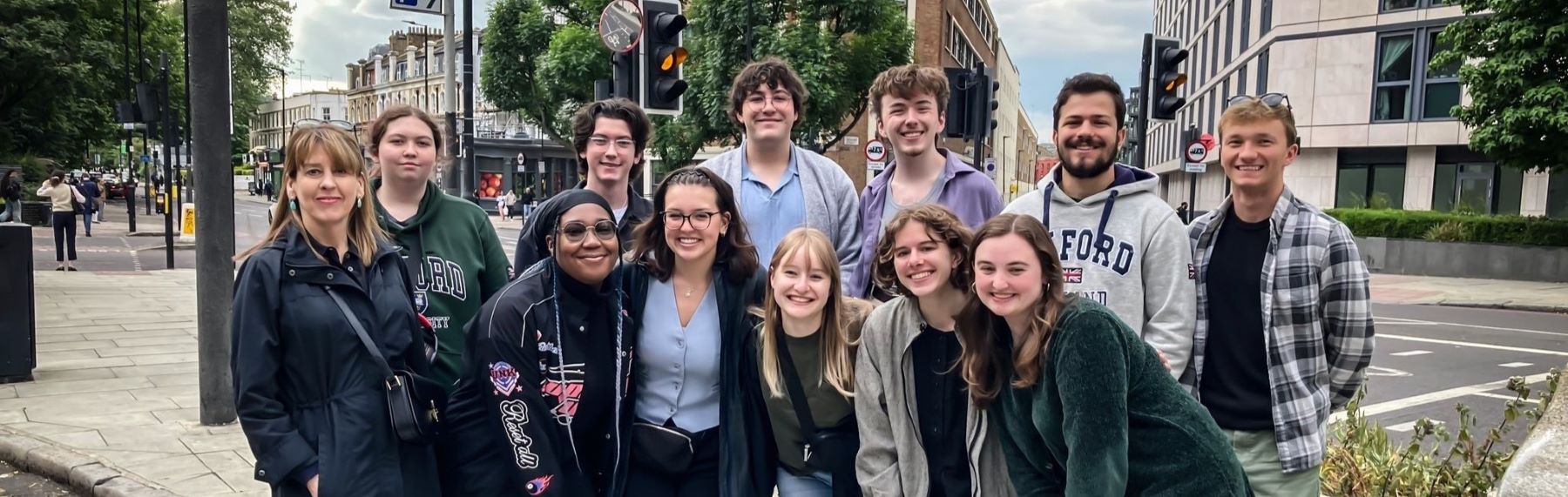 Group of people stand outside on a sidewalk near a street intersection.