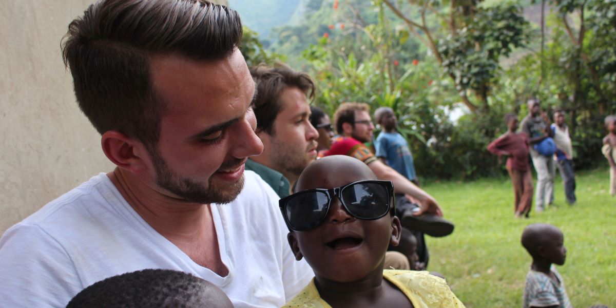 Person with a child wearing oversized sunglasses smiling at the camera with several people standing on a grassy area in the background.