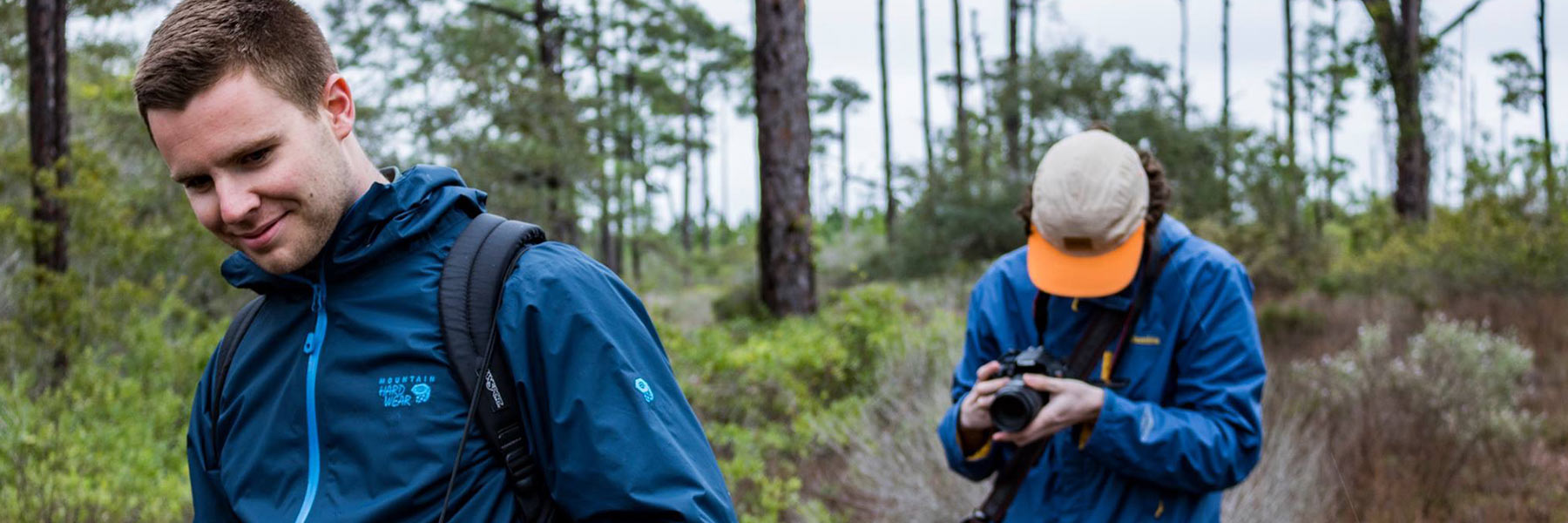 Two men walk through a wooded area with camera equipment.